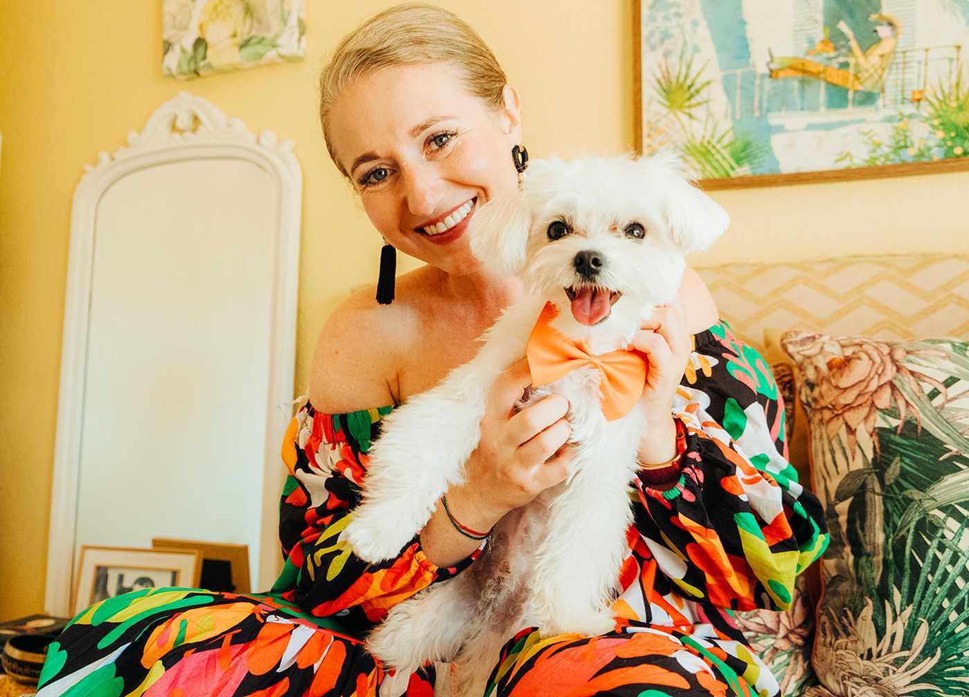 Smiling Marusha Belle in a colorful dress holding a small white dog with an orange bow tie
